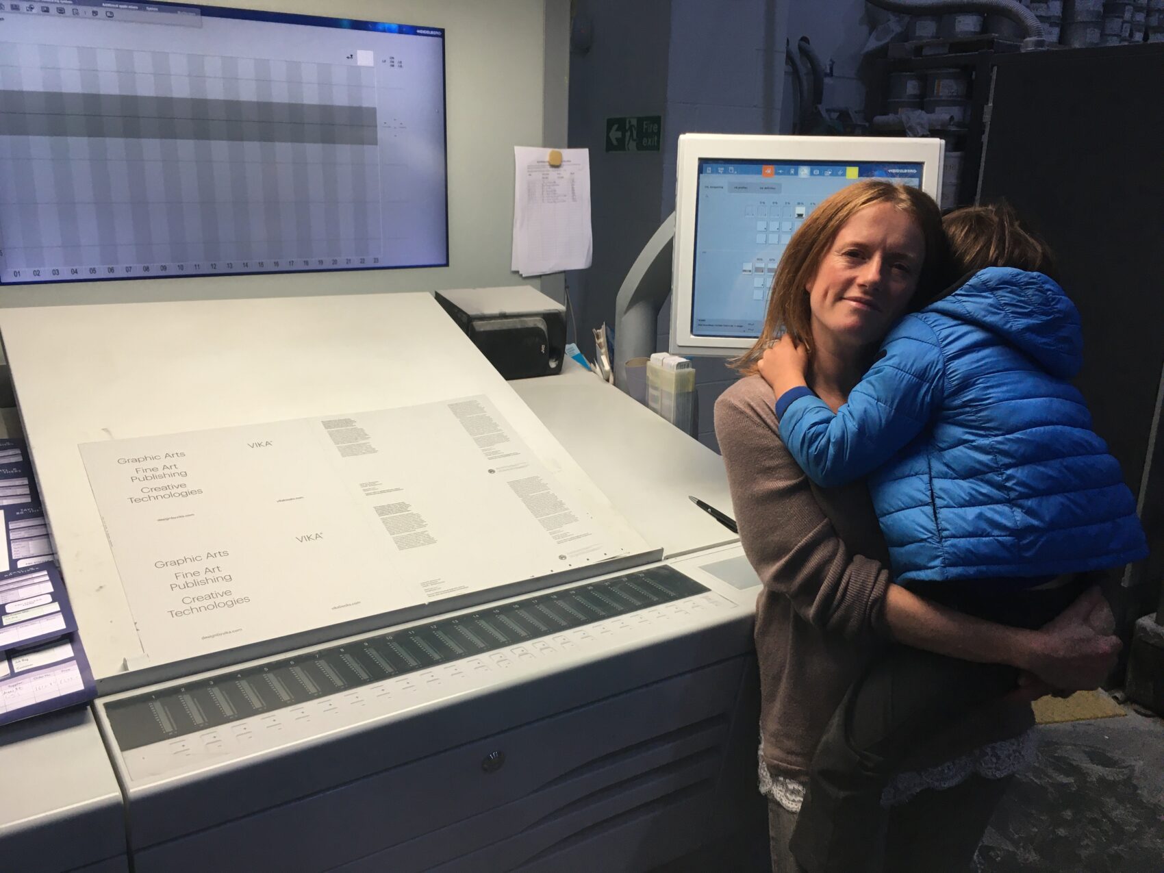 Victoria Forrest, a woman with redhair standing next to the control panel for a commerical lithographic printing press, on which you can see a B2 sheet printed with the words VIKA. Victoria is holding small child in her arms, he is facing away and wearing a blue jacket.